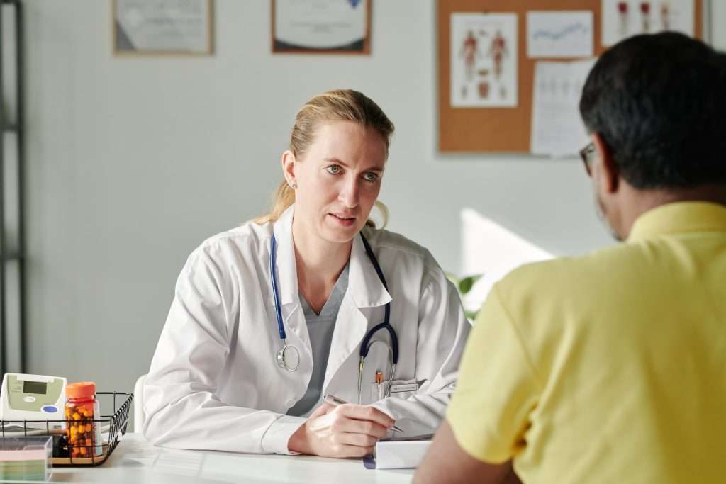 Young female general practitioner in lab coat looking at male patient sitting in front of her and giving him medical advice