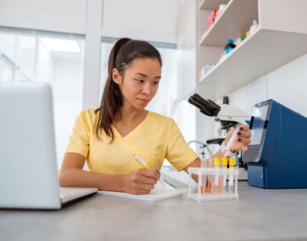Asian lab assistant working with test tubes and taking notes about test results in clinical laboratory